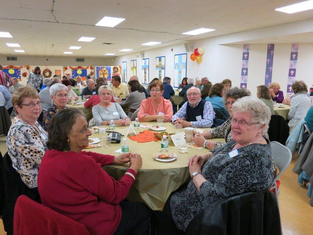 People seated at round tables in a bright community hall, chatting and eating during a social gathering