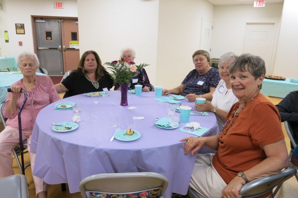 Women seated around a lavender-covered table in a bright dining room, sharing tea and cake