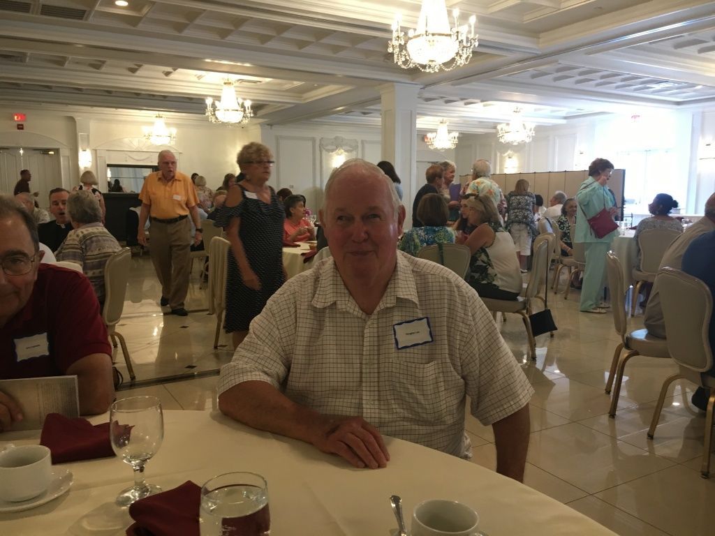 Dining hall with people seated at round tables under chandeliers; man in plaid shirt sits in foreground.
