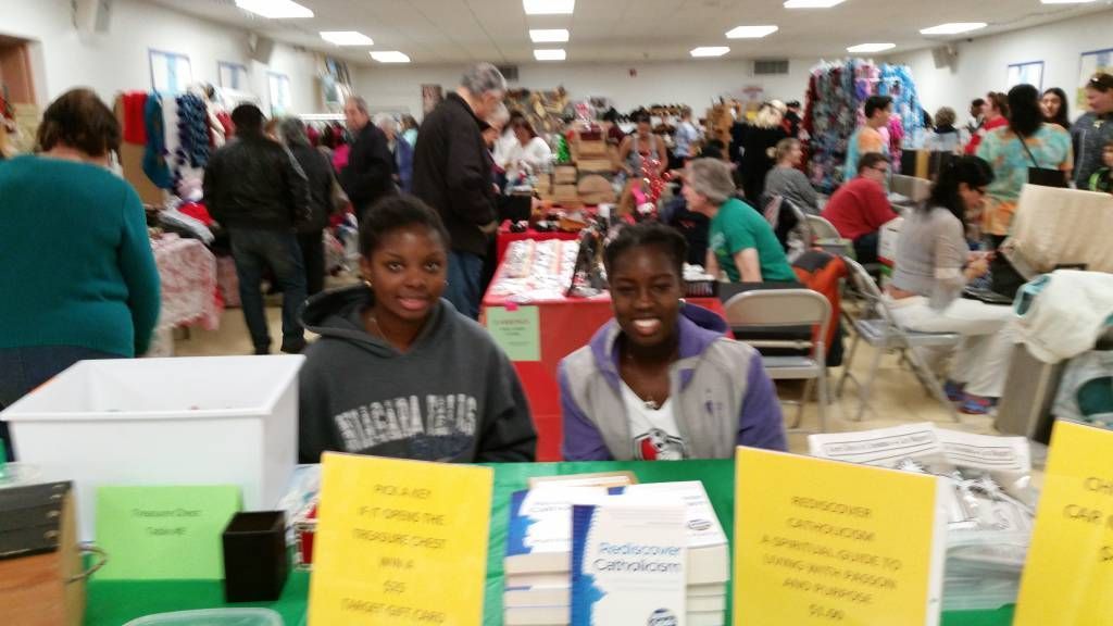 Two people smiling at a table in a busy indoor community event with signs and boxes in front.