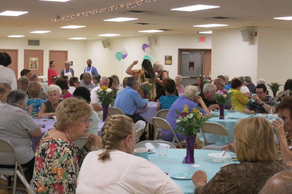 Crowded community banquet hall with guests seated at round tables, colorful balloons, and floral centerpieces