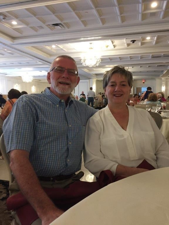 Two people smiling at a banquet table in a bright ballroom with chandeliers