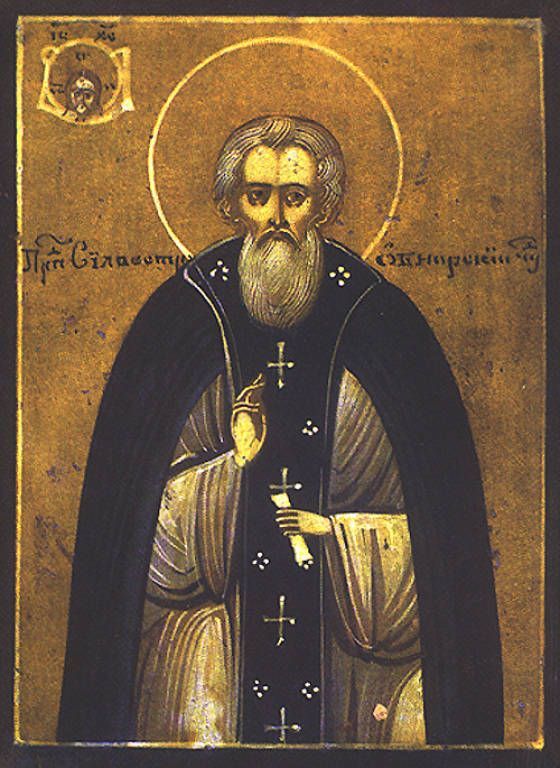 Byzantine icon of a bearded saint in dark robes holding a cross and blessing gesture on a gold background