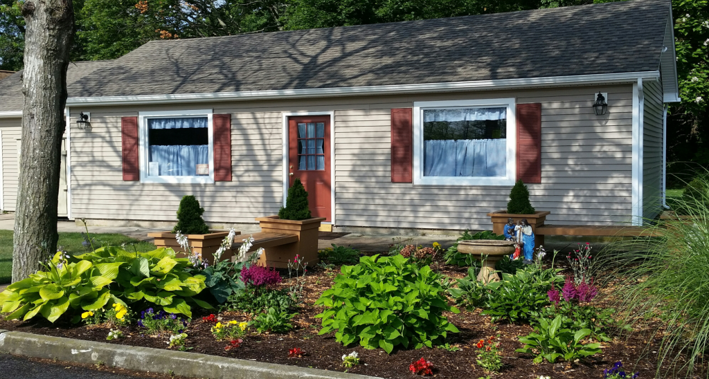 Small beige house with white shutters and a colorful front garden under trees