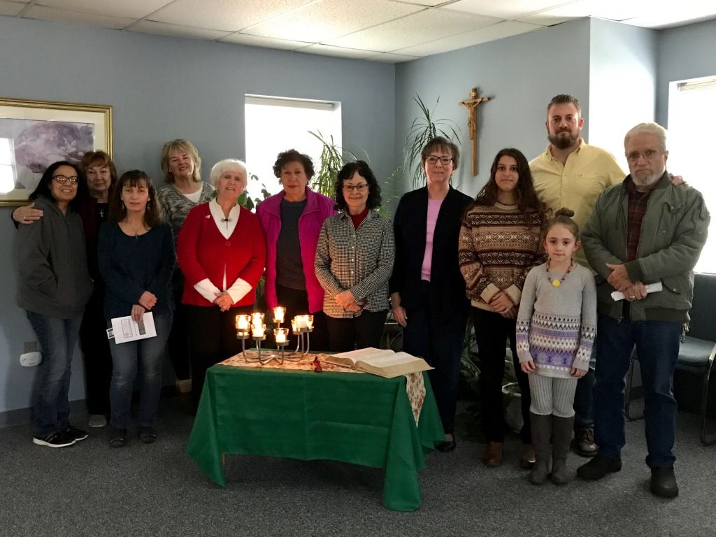 Group of people standing around a candlelit table in a small room, gathered for a ceremony or celebration.