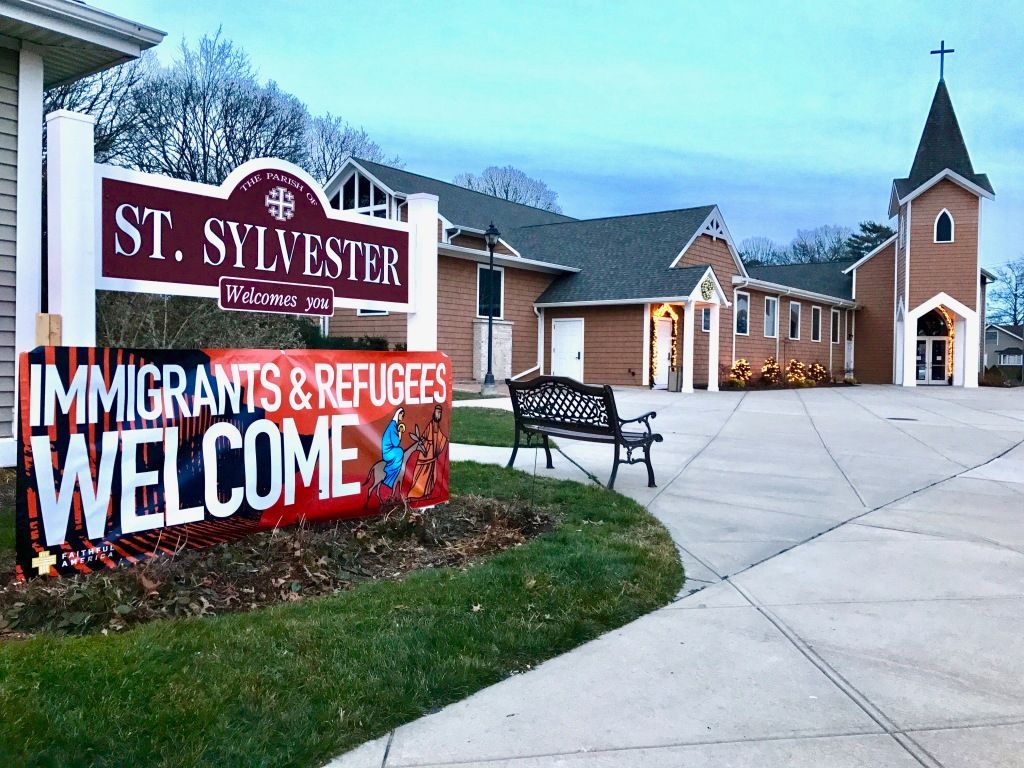 St. Sylvester church sign reading “Immigrants & Refugees Welcome” beside a church and walkway