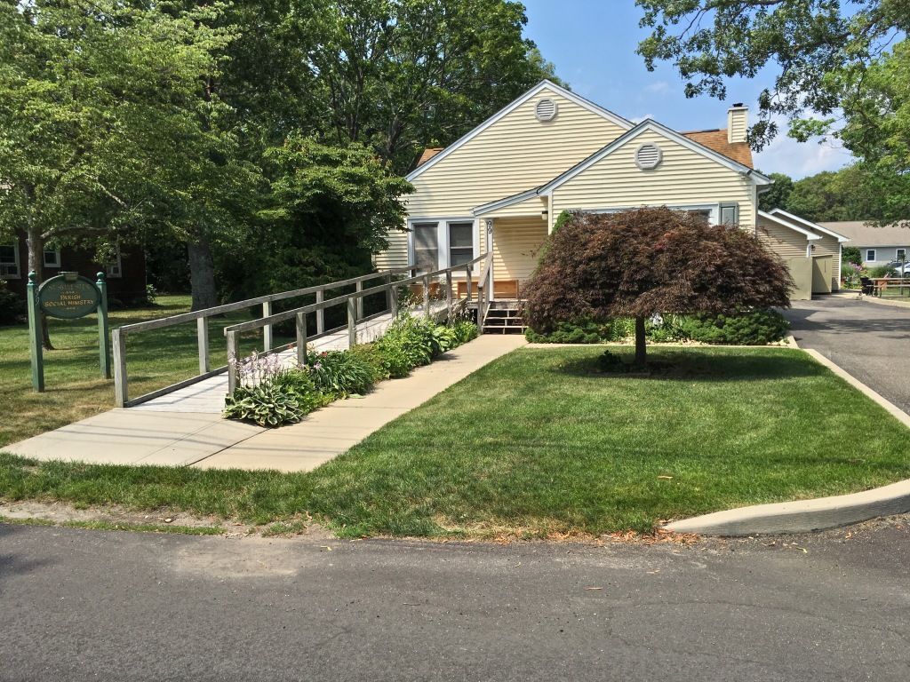 Small yellow house with wheelchair ramp, flower beds, and green lawn on a sunny street corner
