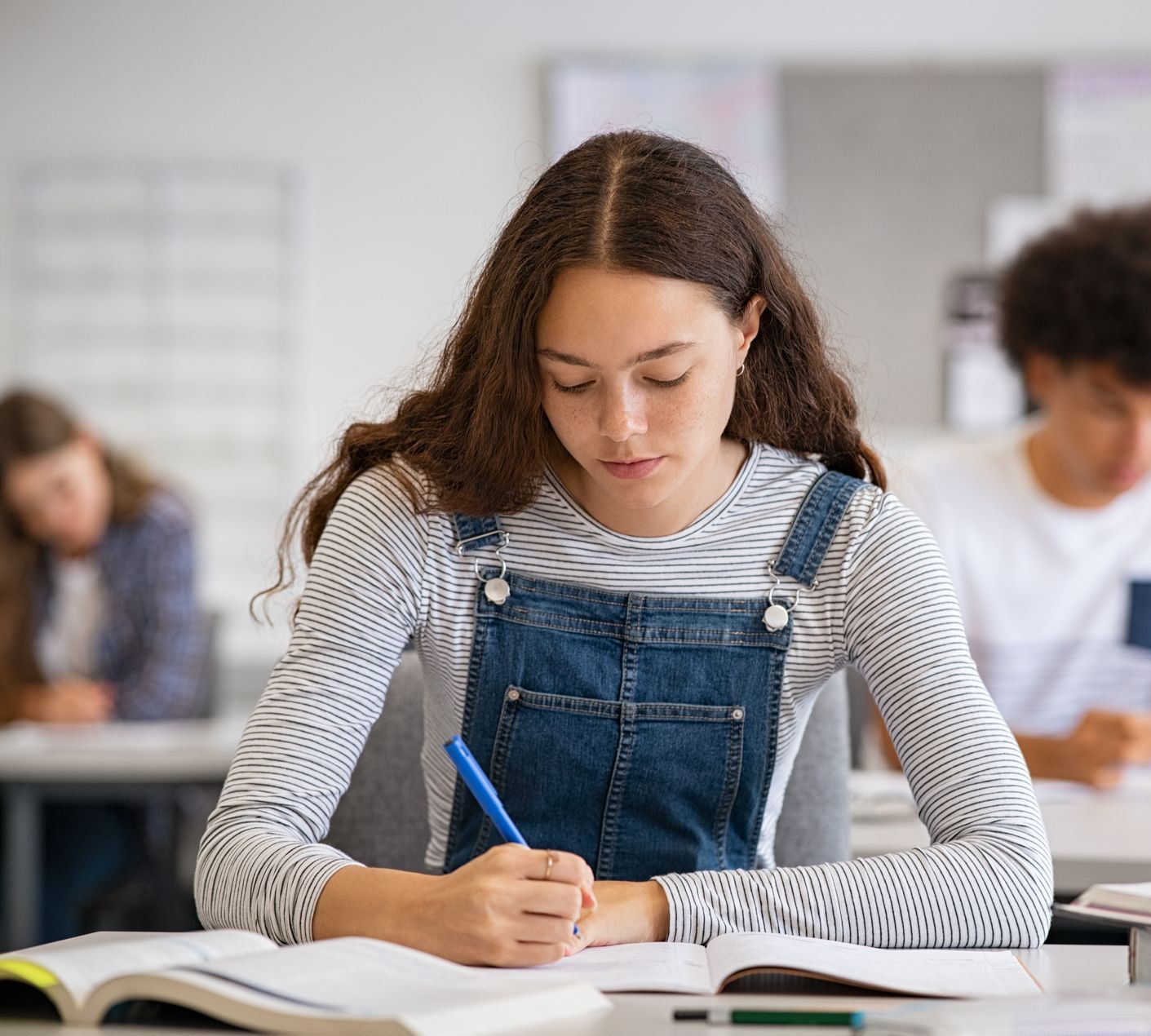 A young woman is sitting at a desk in a classroom writing in a notebook.