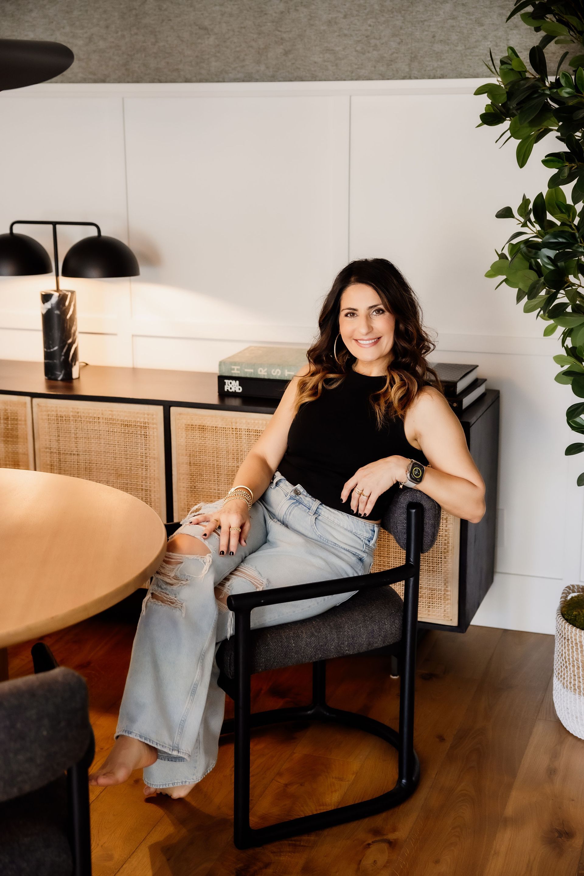 Mania Persekian, REALTOR, is seated and smiling in a modern-styled room in a black top and jeans.
