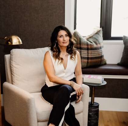 Mania Persekian, REALTOR, seated on a sofa in a warmly lit professional brand photo.
