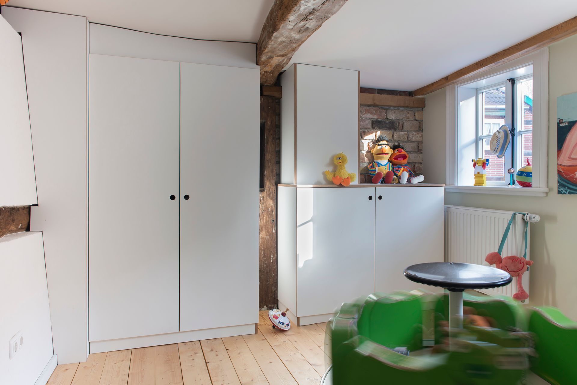 Helles Dachzimmer mit weißen Schränken, freiliegenden Balken, einem Fenster und einem kleinen Tisch mit grünen Stühlen.