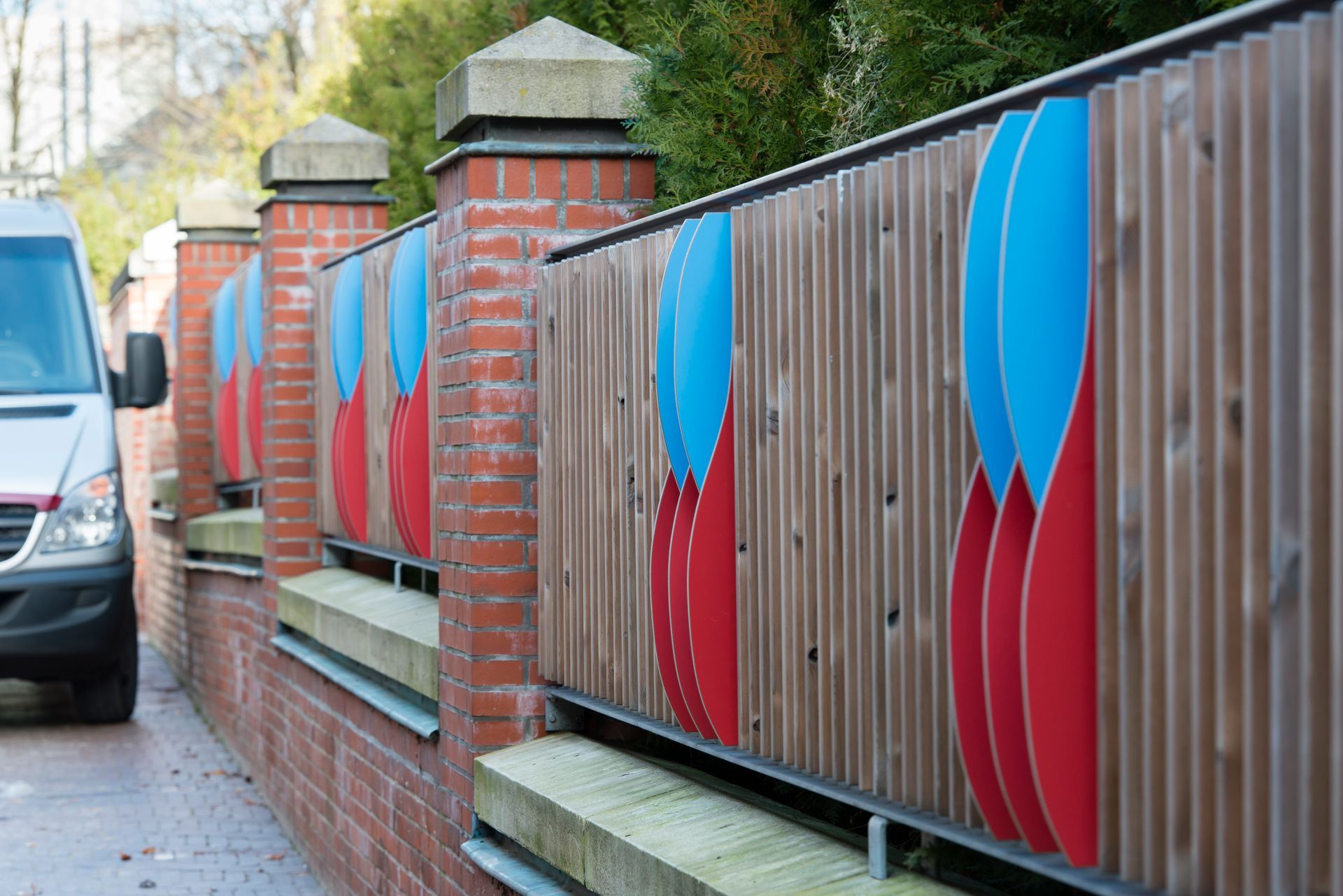 Eine Reihe roter und blauer Paneele an einem Zaun aus Ziegeln und Holz neben einem Gehweg und einem geparkten Lieferwagen.