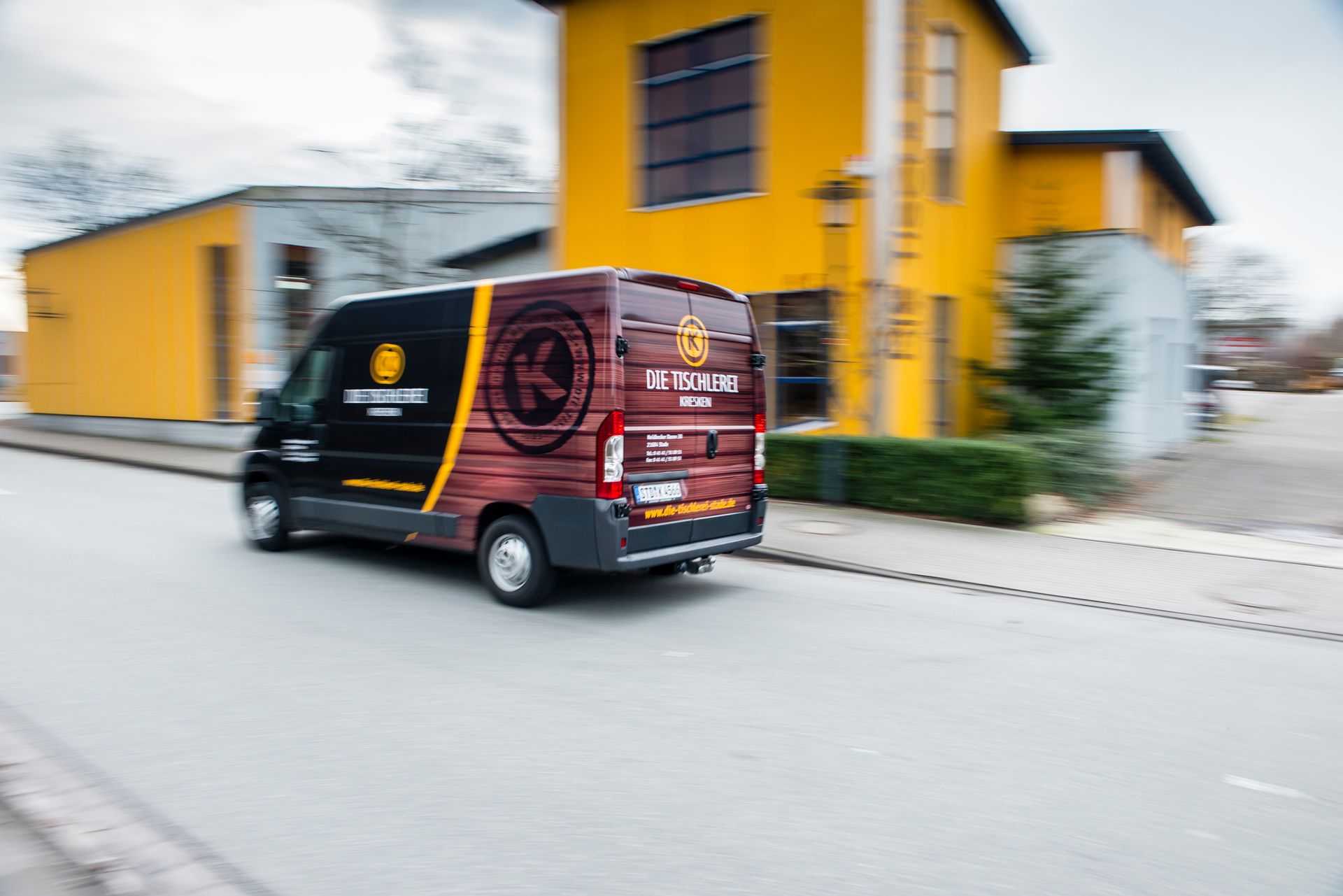 Ein schwarzer Lieferwagen mit roter Grafik fährt an leuchtend gelben Gebäuden auf einer Straße vorbei.