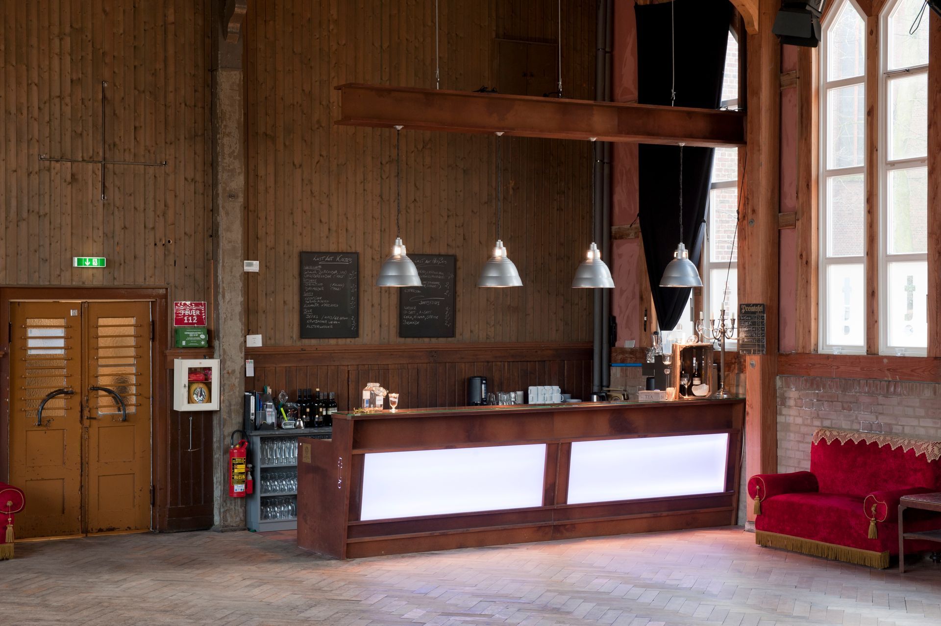 Holzgetäfelte Hotellobby mit Rezeption, Hängelampen, großen Fenstern und roten Sofas.