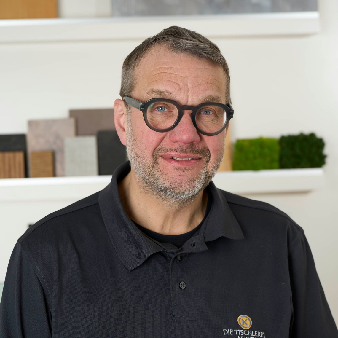 Porträt eines lächelnden Mannes mit Brille in einem schwarzen Polohemd, der in einem Innenraum steht, hinter ihm Regale.