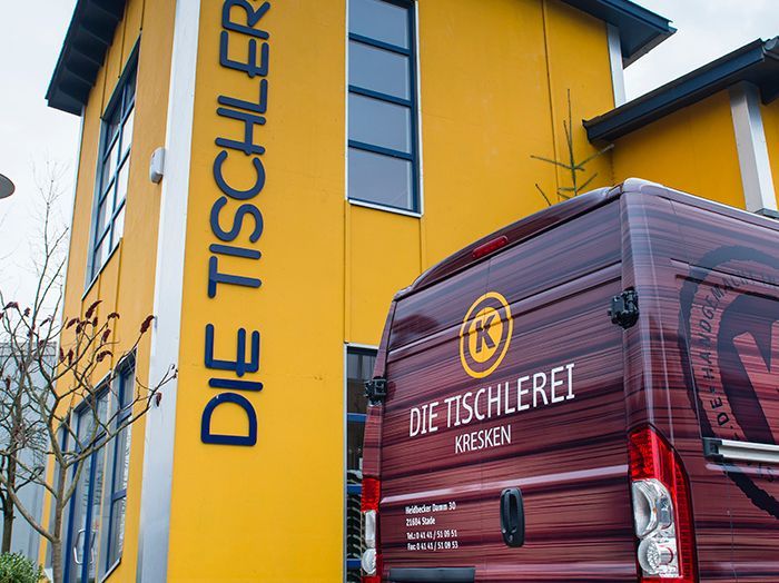 Gelbes Gebäude mit dem Schild „DIE TISCHLEREI“ und rotem Lieferwagen davor