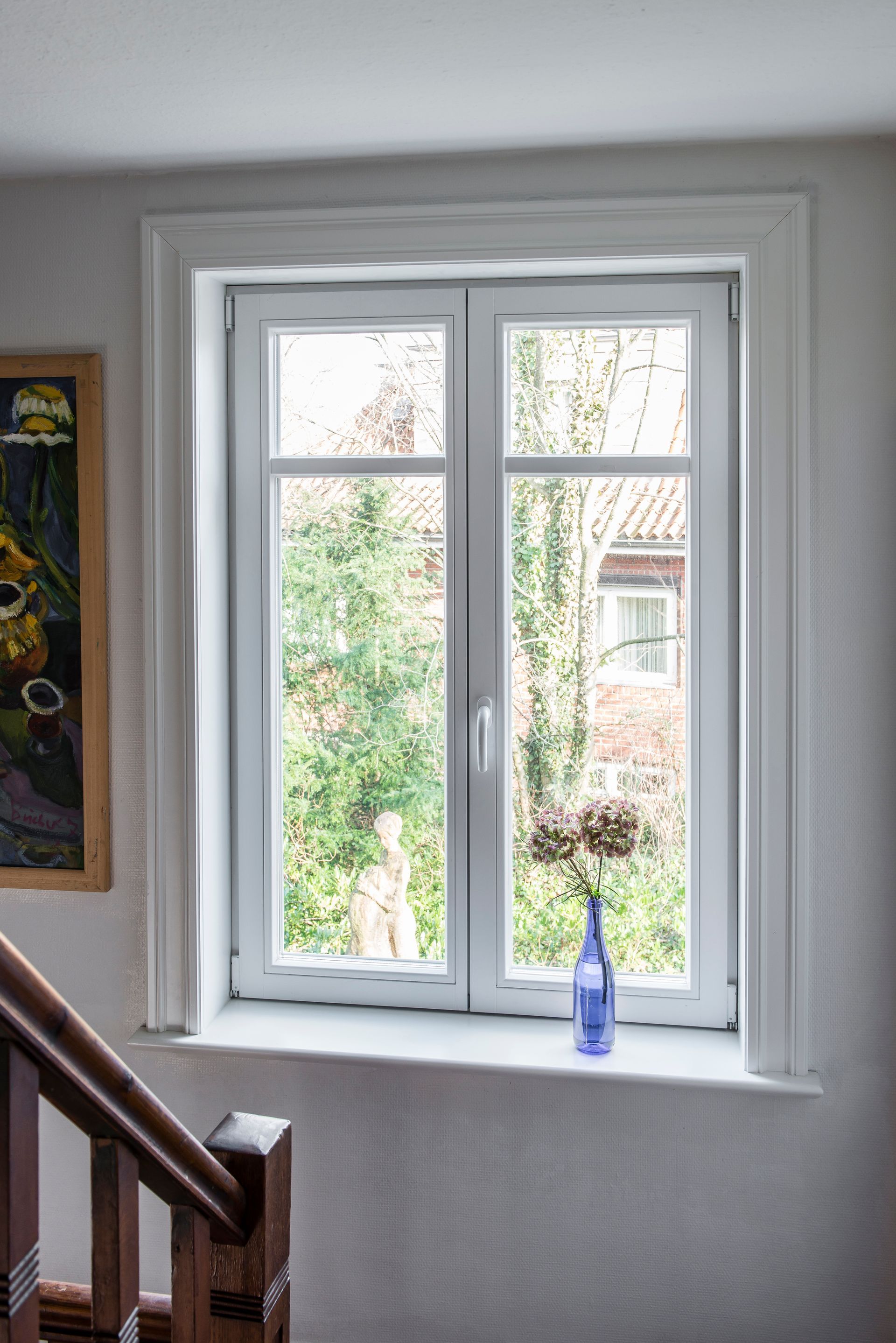 Ein weiß gerahmtes Fenster mit Grünpflanzen draußen und einer blauen Flasche auf dem Fensterbrett neben einer Treppe