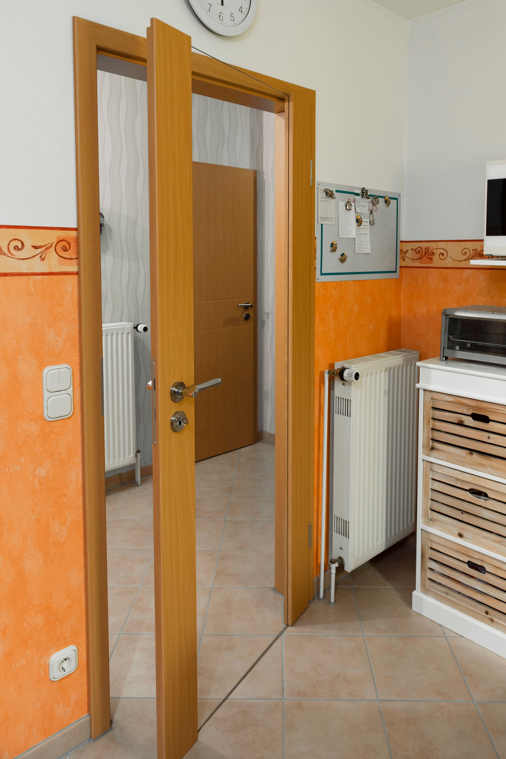 Offene Holztür in einem gefliesten Flur mit orangefarbenen Wänden, Heizkörper und einer kleinen Theke.