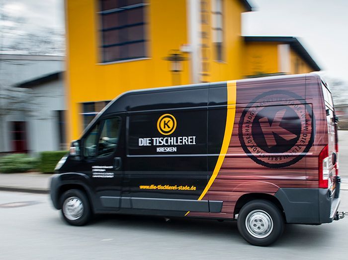 Schwarzer Lieferwagen mit der Aufschrift „Die Tischlerei“ fährt an einem gelben Gebäude vorbei.