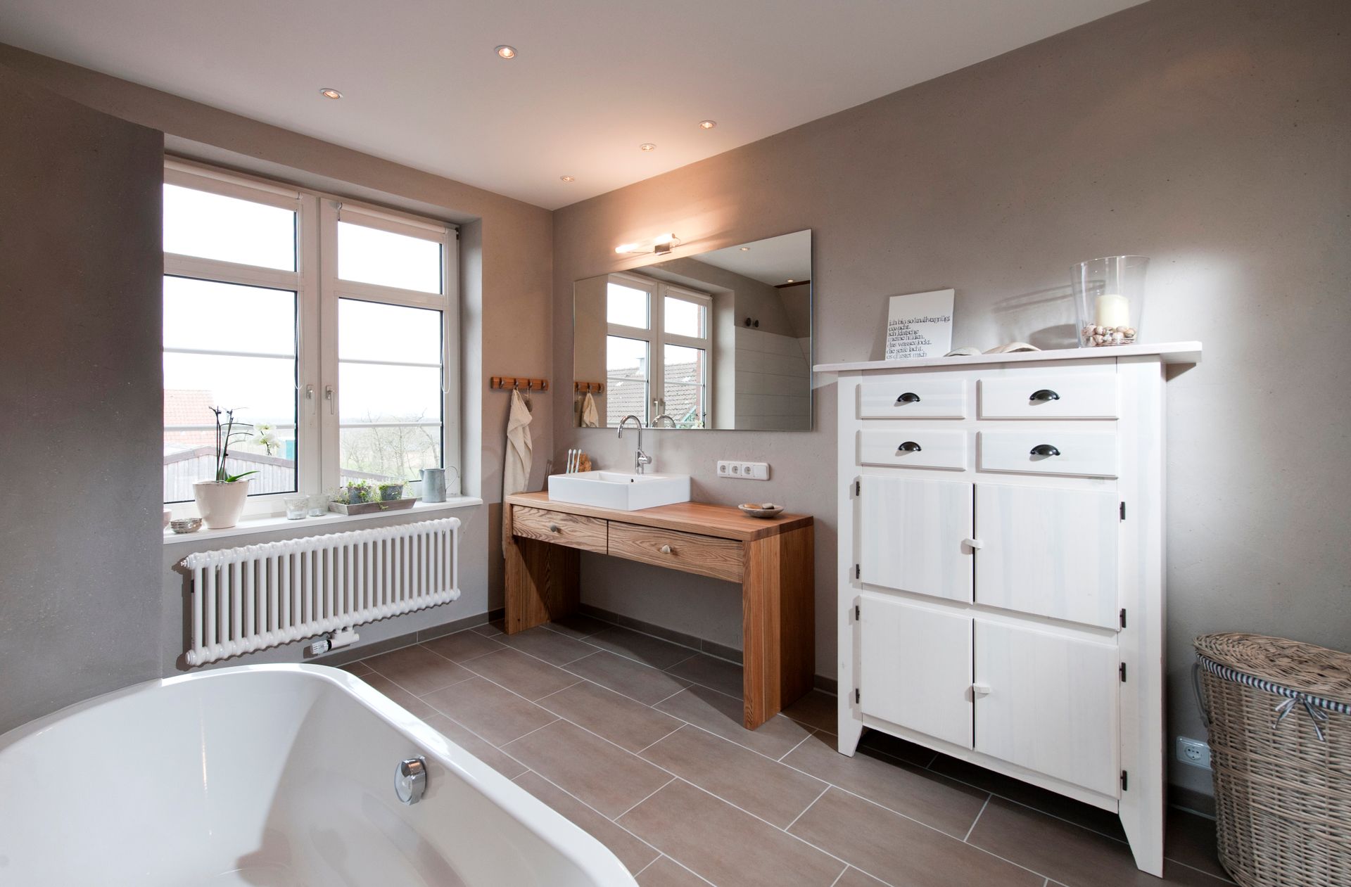 Helles Badezimmer mit freistehender Badewanne, Waschtisch aus Holz, weißem Schrank und großem Fenster.