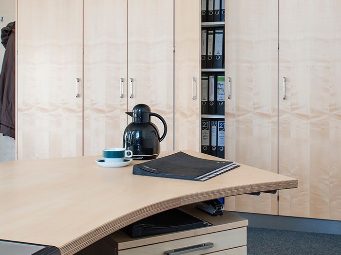 Holztisch in einem modernen Büro, mit Tasse, Karaffe und Notizbuch vor Schränken.