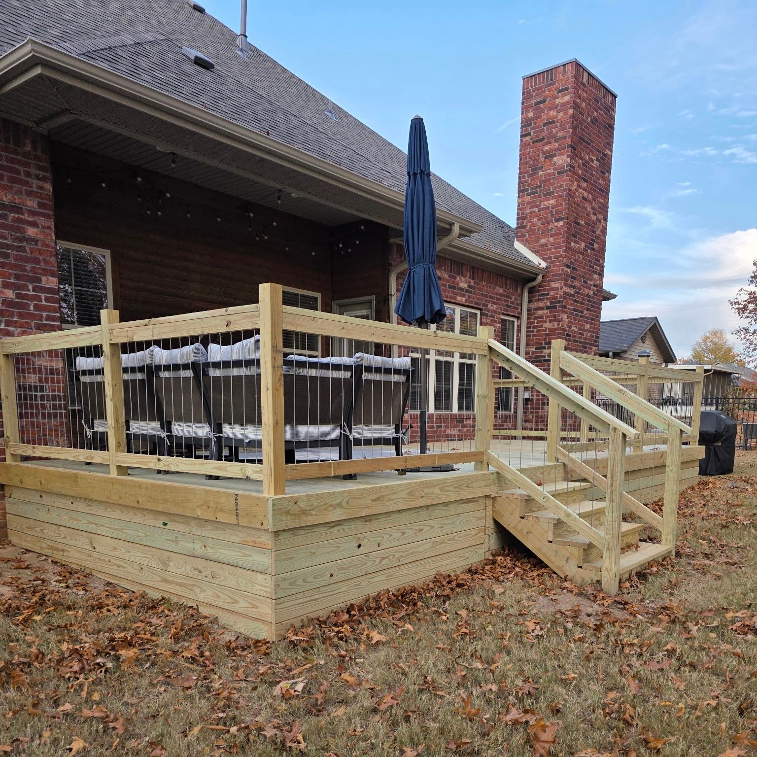 Wooden backyard deck with railings, stairs, outdoor dining set, and closed umbrella beside a brick house.
