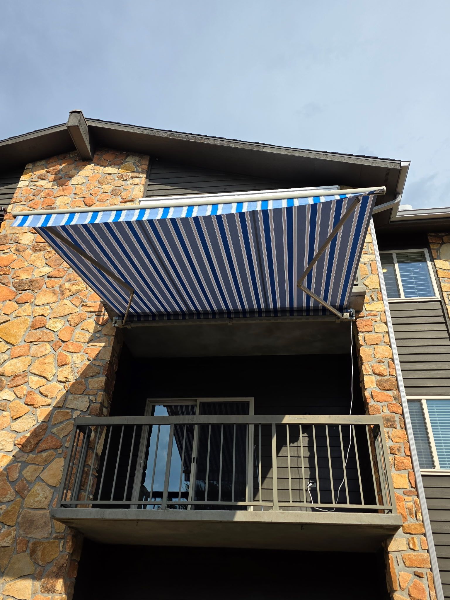 Stone apartment balcony with blue-and-white striped awning and black railing under a cloudy sky