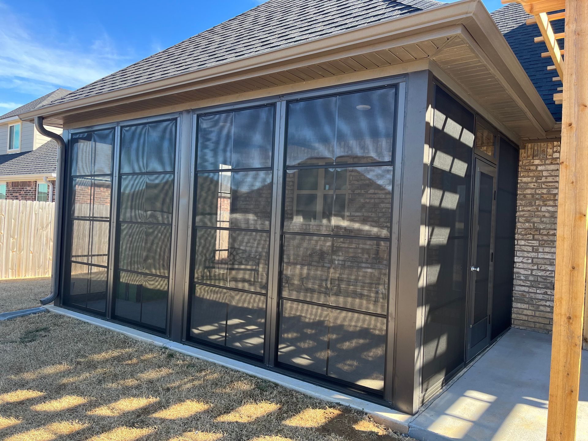 Black-framed screened patio enclosure beside a brick house in a sunny backyard
