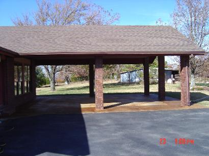Covered outdoor pavilion with brick posts and a shingled roof beside a paved lot, with trees in the background