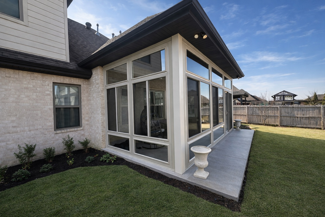 Glass-enclosed sunroom attached to a house, with a small patio and grassy yard.