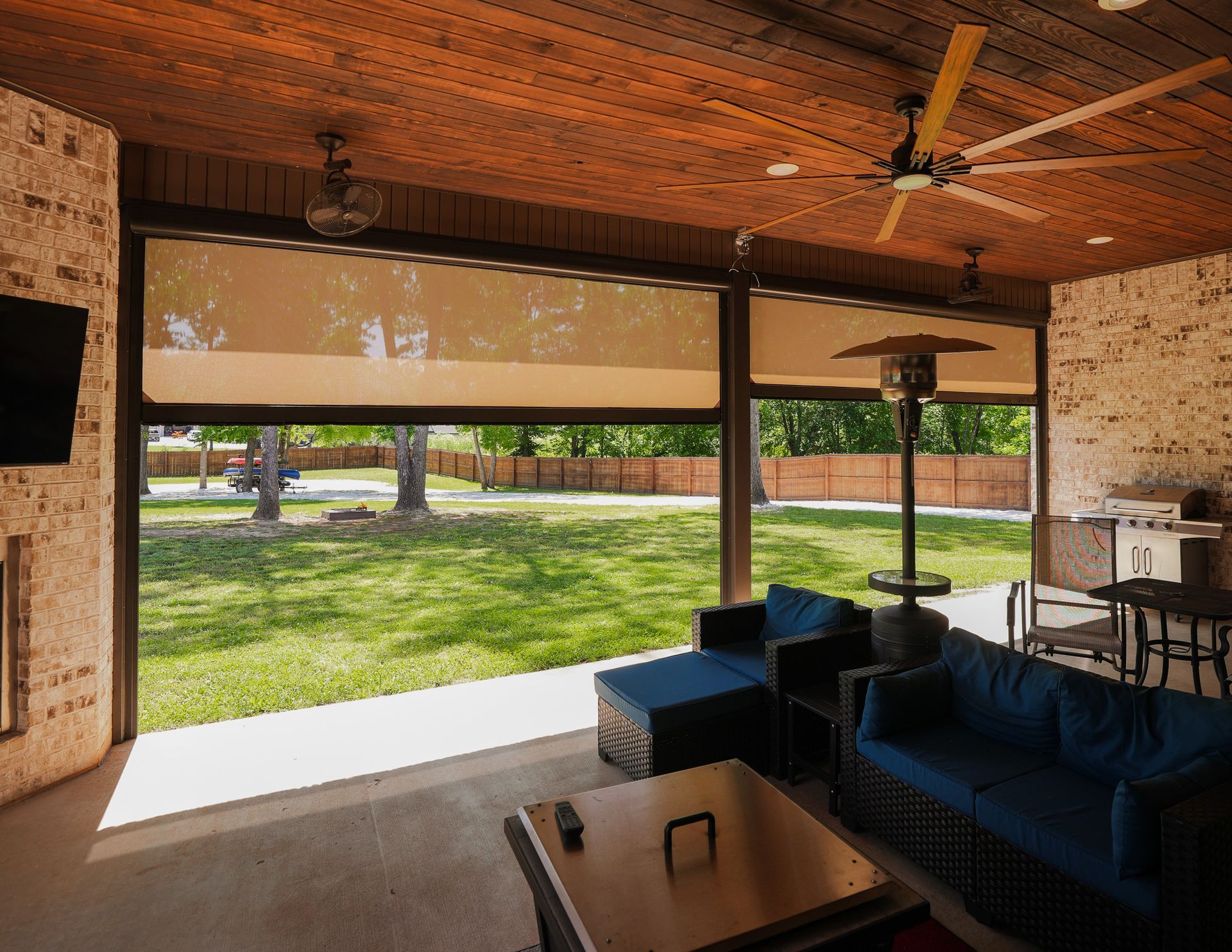 Covered patio with sectional sofa, ceiling fan, table, and open view of a grassy yard and trees.
