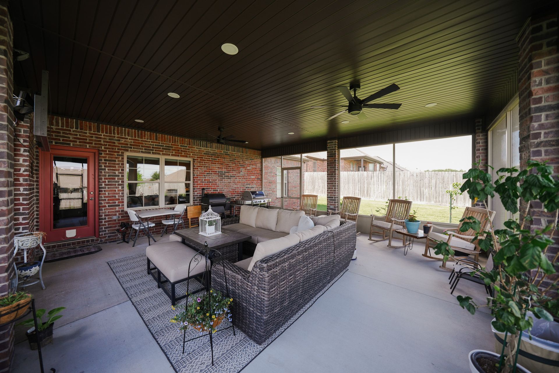 Covered patio with wicker seating, brick walls, potted plants, and a ceiling fan overlooking a backyard.