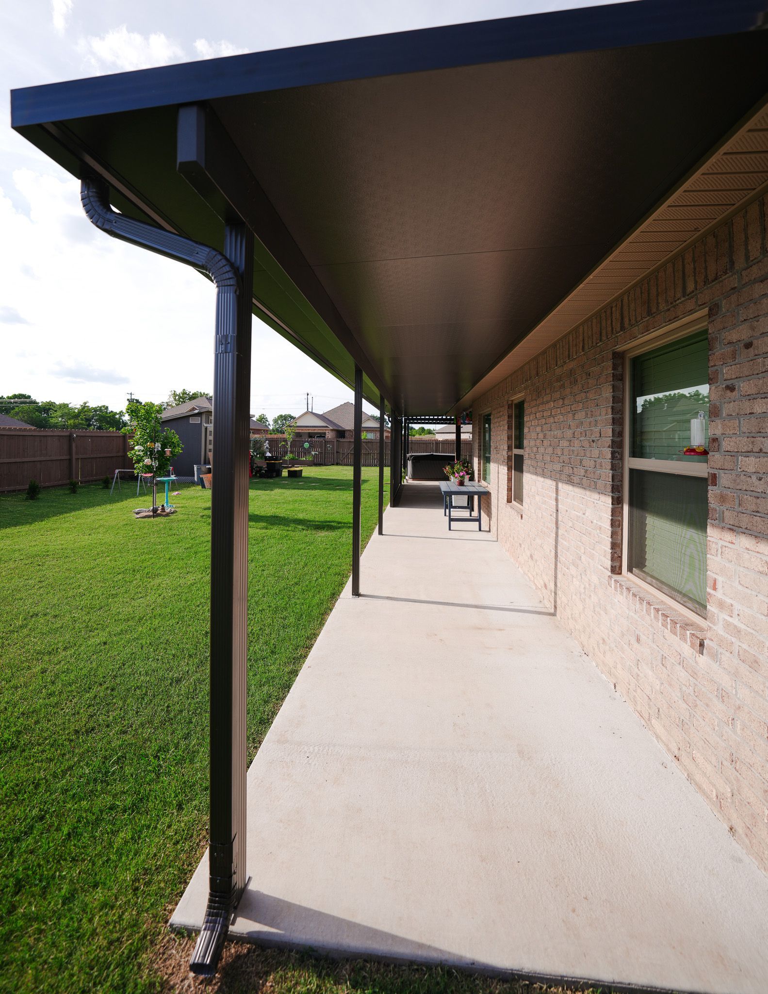 Covered concrete walkway beside a brick house, with black posts and a green lawn.