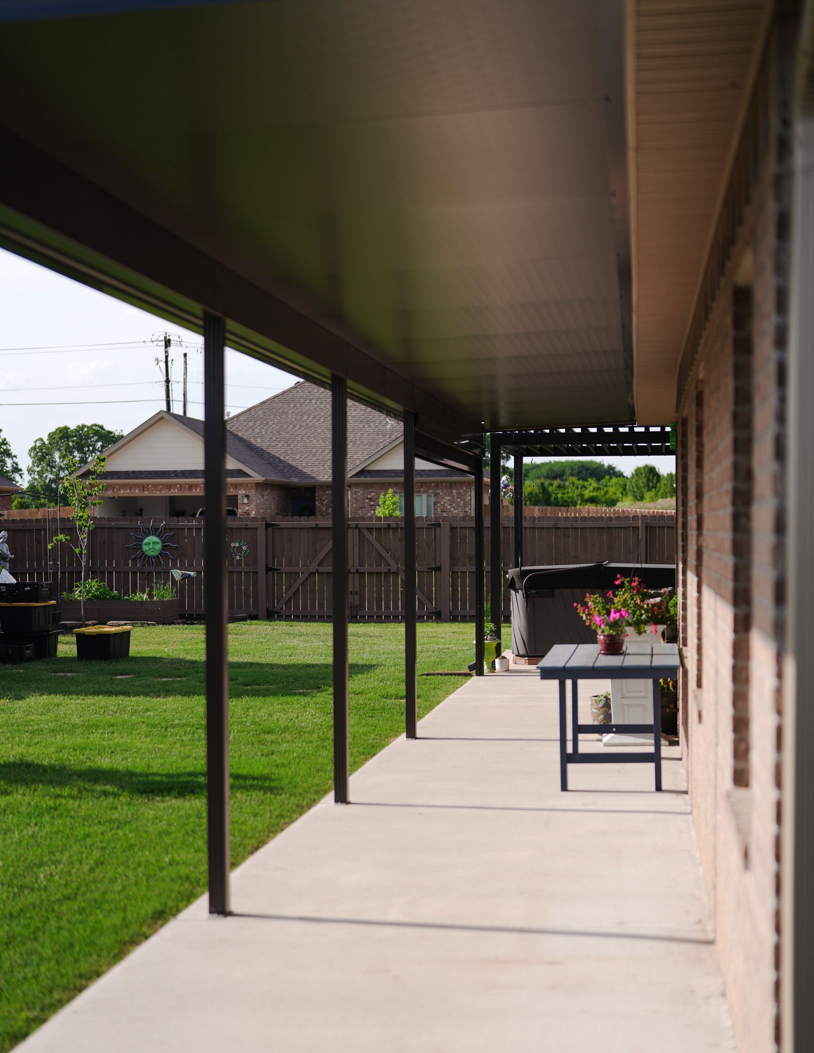Covered backyard patio beside a brick house, with concrete walkway, green lawn, and a small table with flowers.