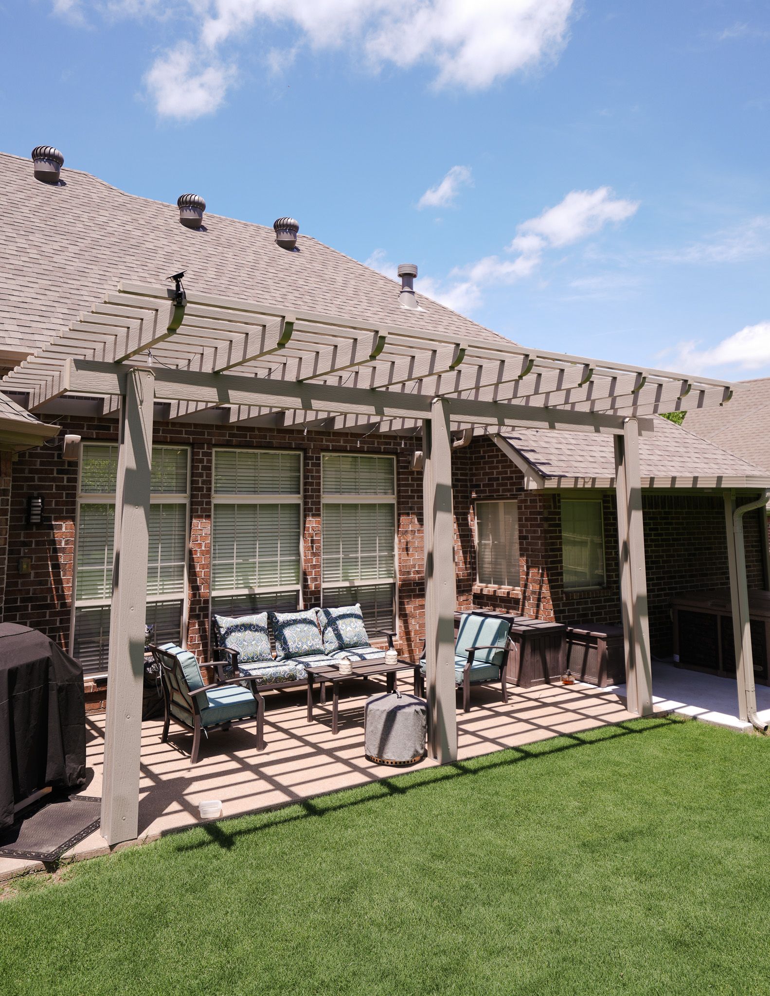 Sunny backyard patio with pergola, cushioned seating, and green lawn beside the house