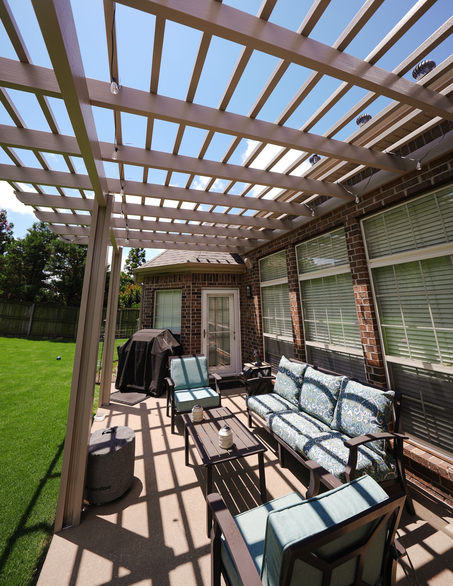 Patio with pergola beams, outdoor sofa, grill, and grassy yard in sunlight