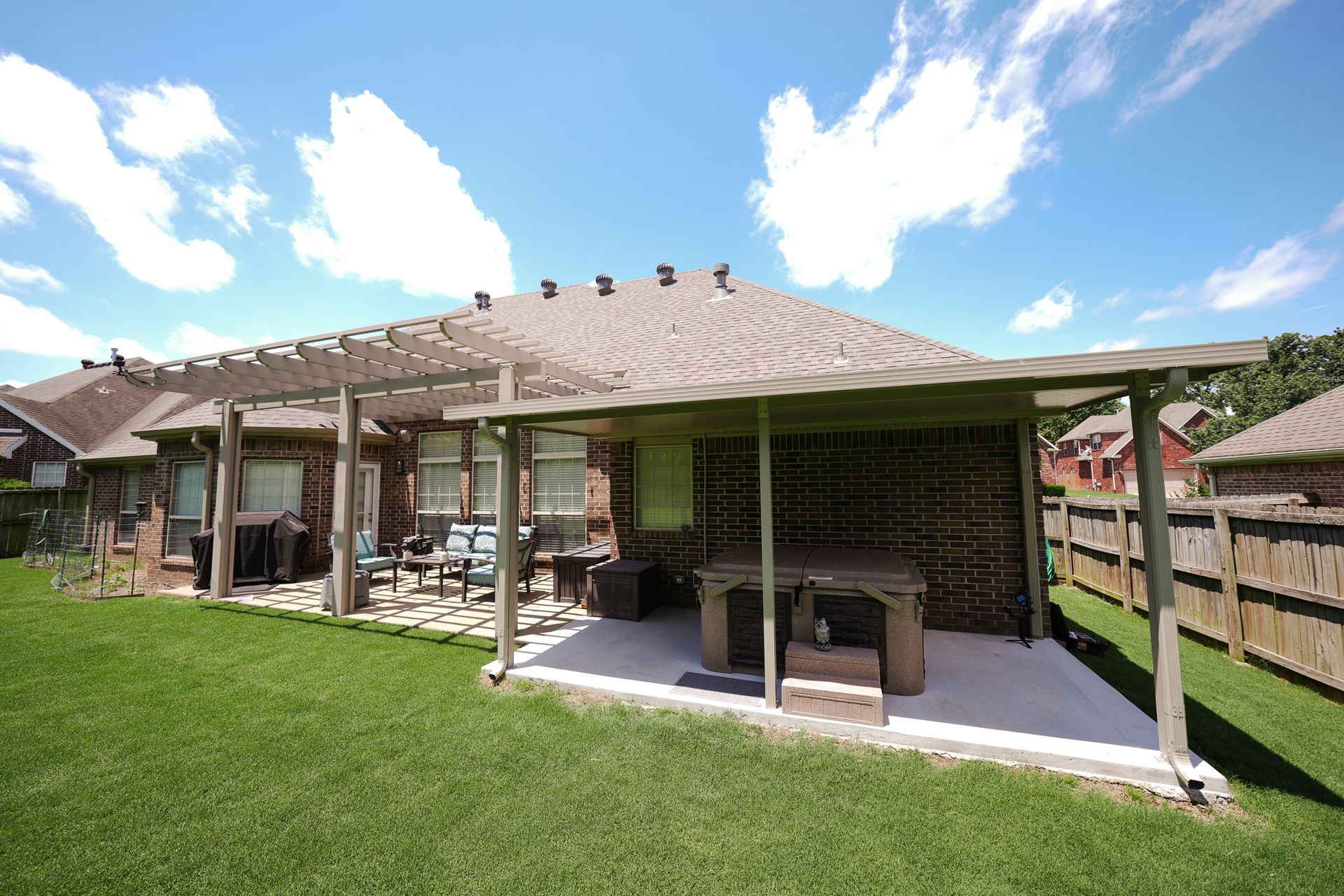 Backyard patio with a covered pergola, outdoor seating, grill, and green lawn under a blue sky