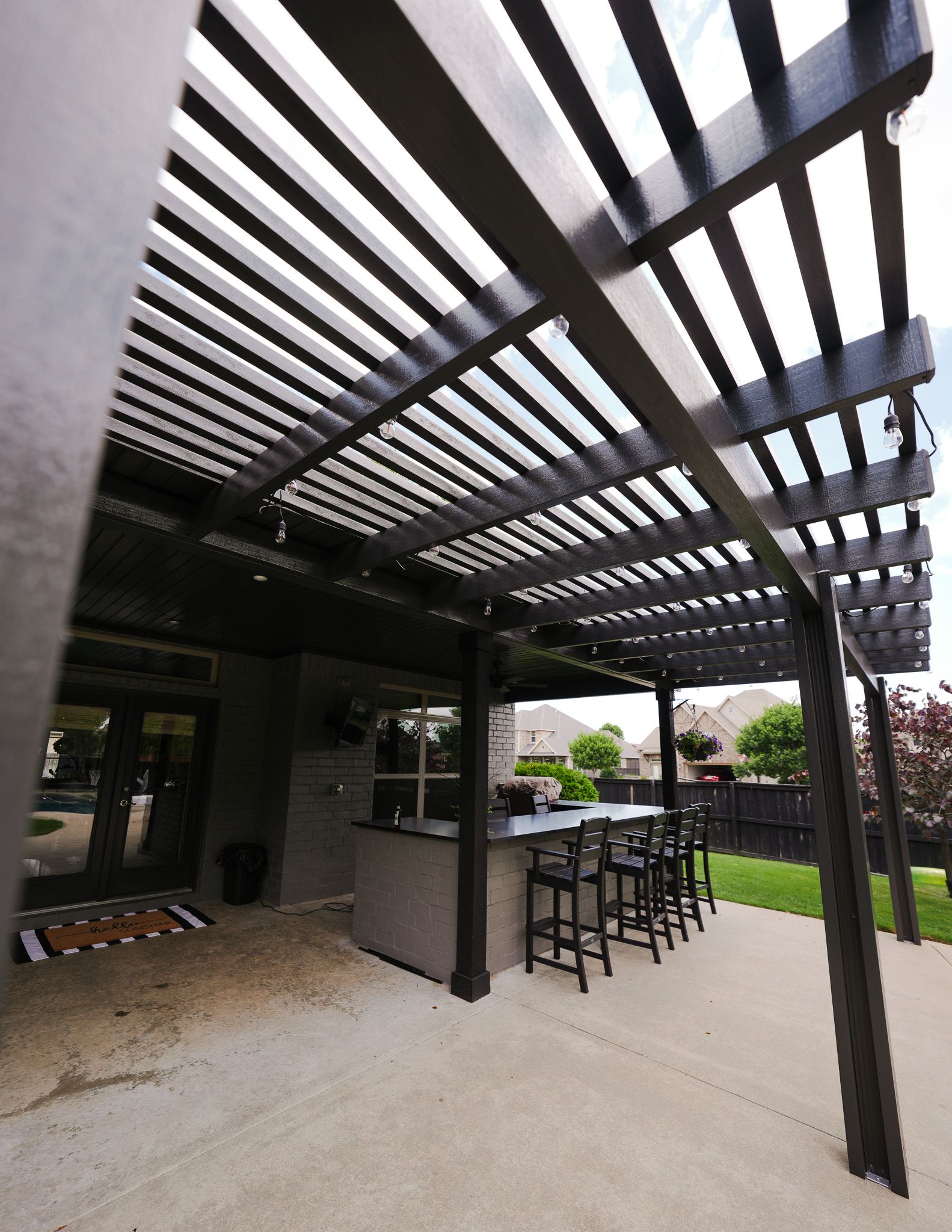 Covered outdoor patio with pergola slats, bar stools, grill, and lawn beyond
