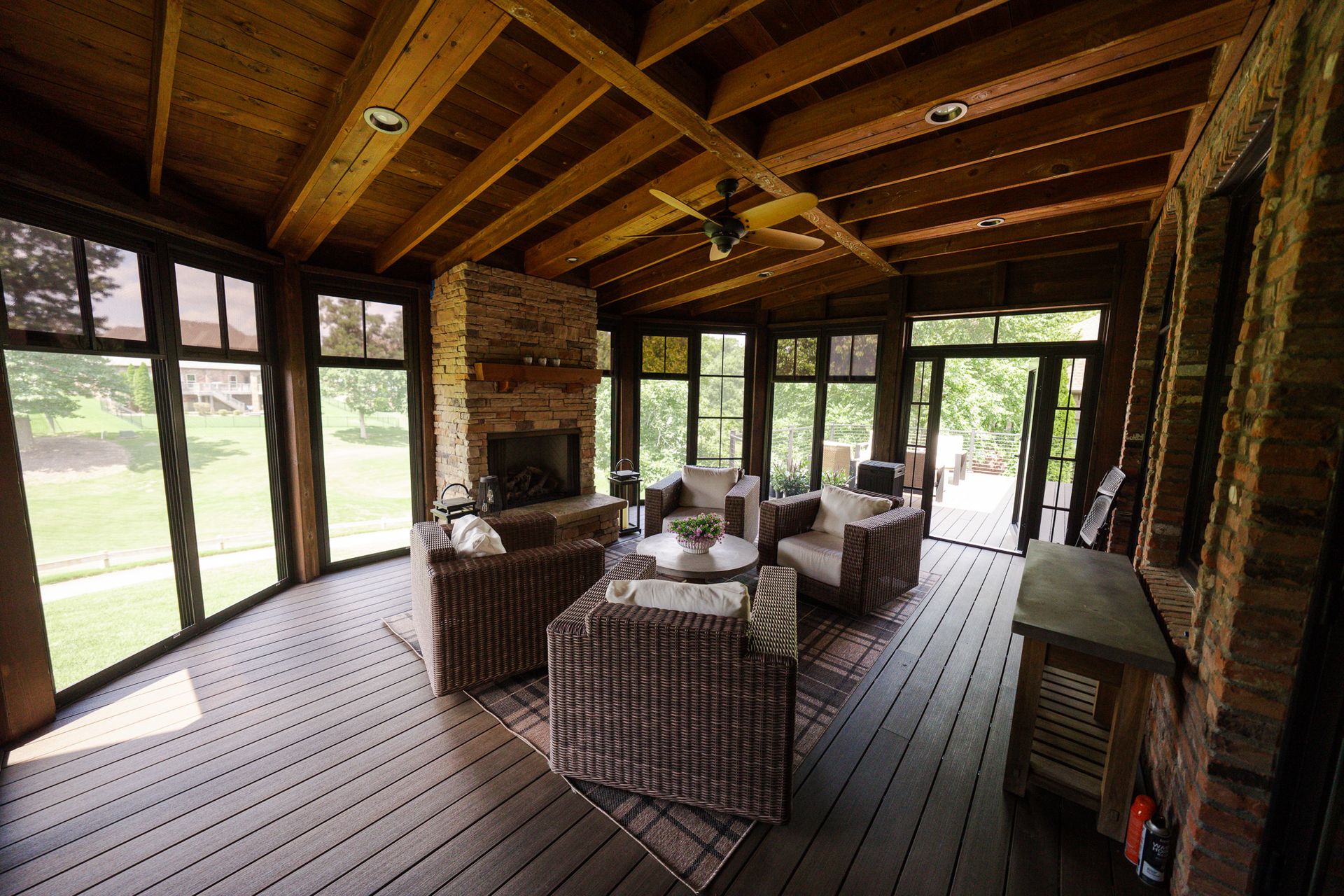 Covered porch with wicker seating, wooden ceiling beams, and large windows overlooking a grassy yard