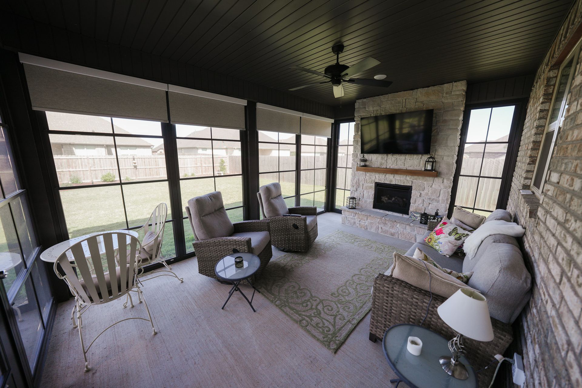 Covered patio with wicker seating, small tables, rug, and a stone fireplace overlooking a yard.