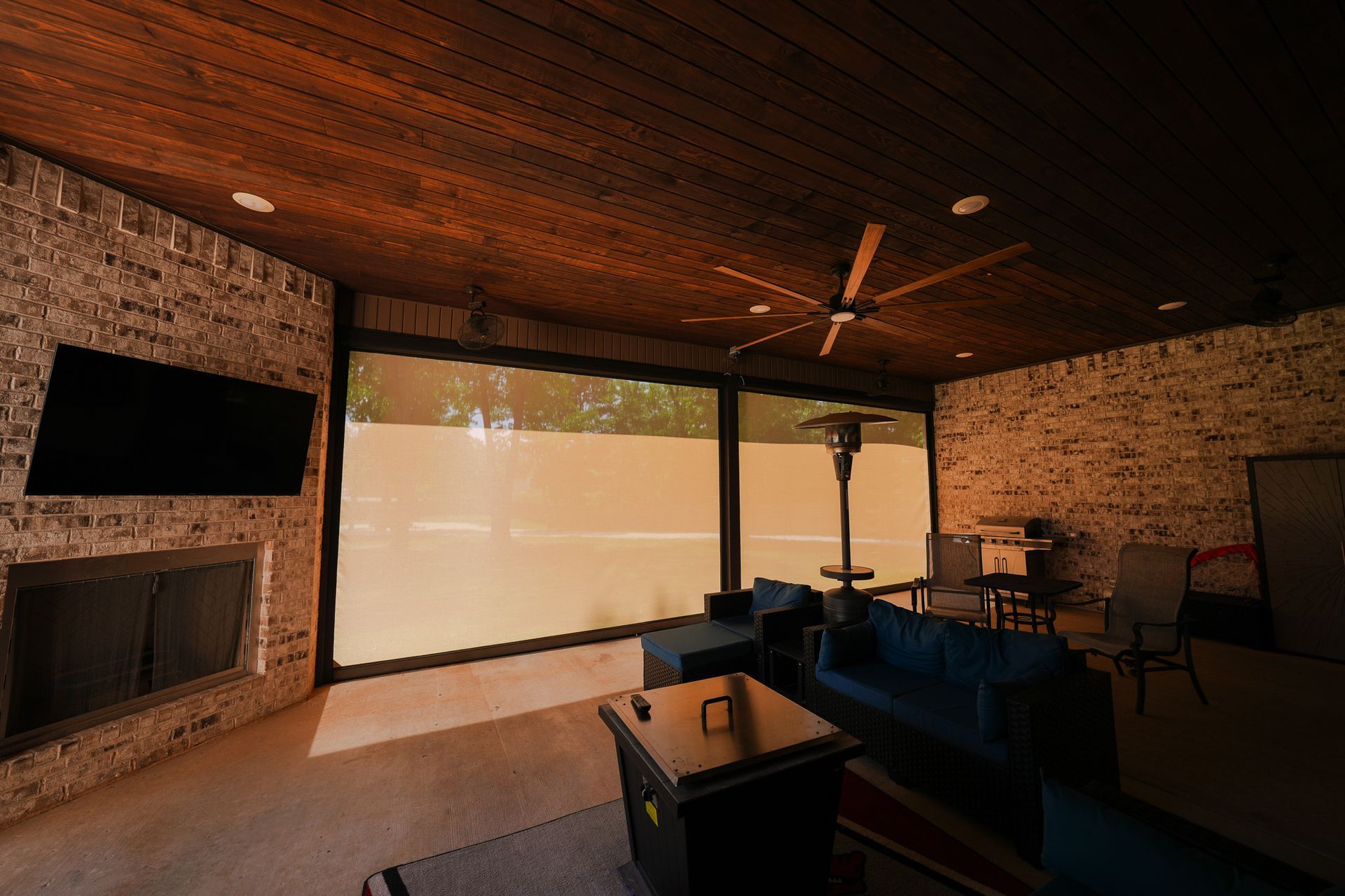 Modern living room with brick walls, dark wood ceiling, large windows, TV, fireplace, and blue sofa