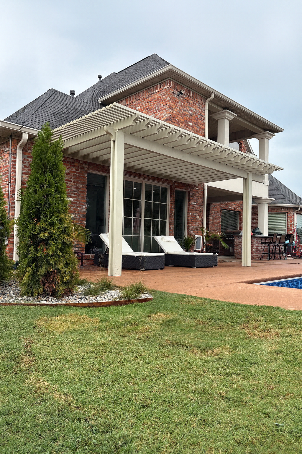 Brick house with covered patio, white columns, lounge chairs, and a swimming pool beside a lawn.