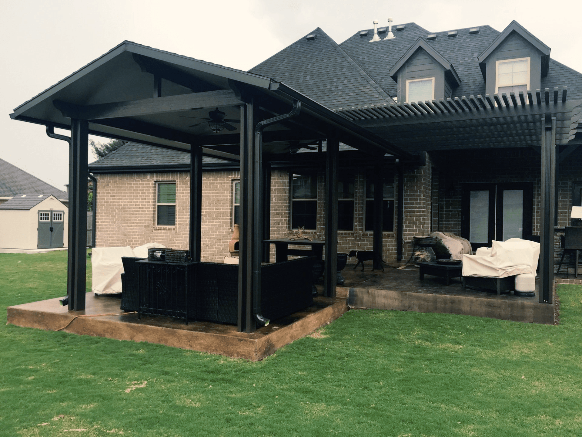 Covered backyard patio with black metal frame, brick house, outdoor seating, and green lawn