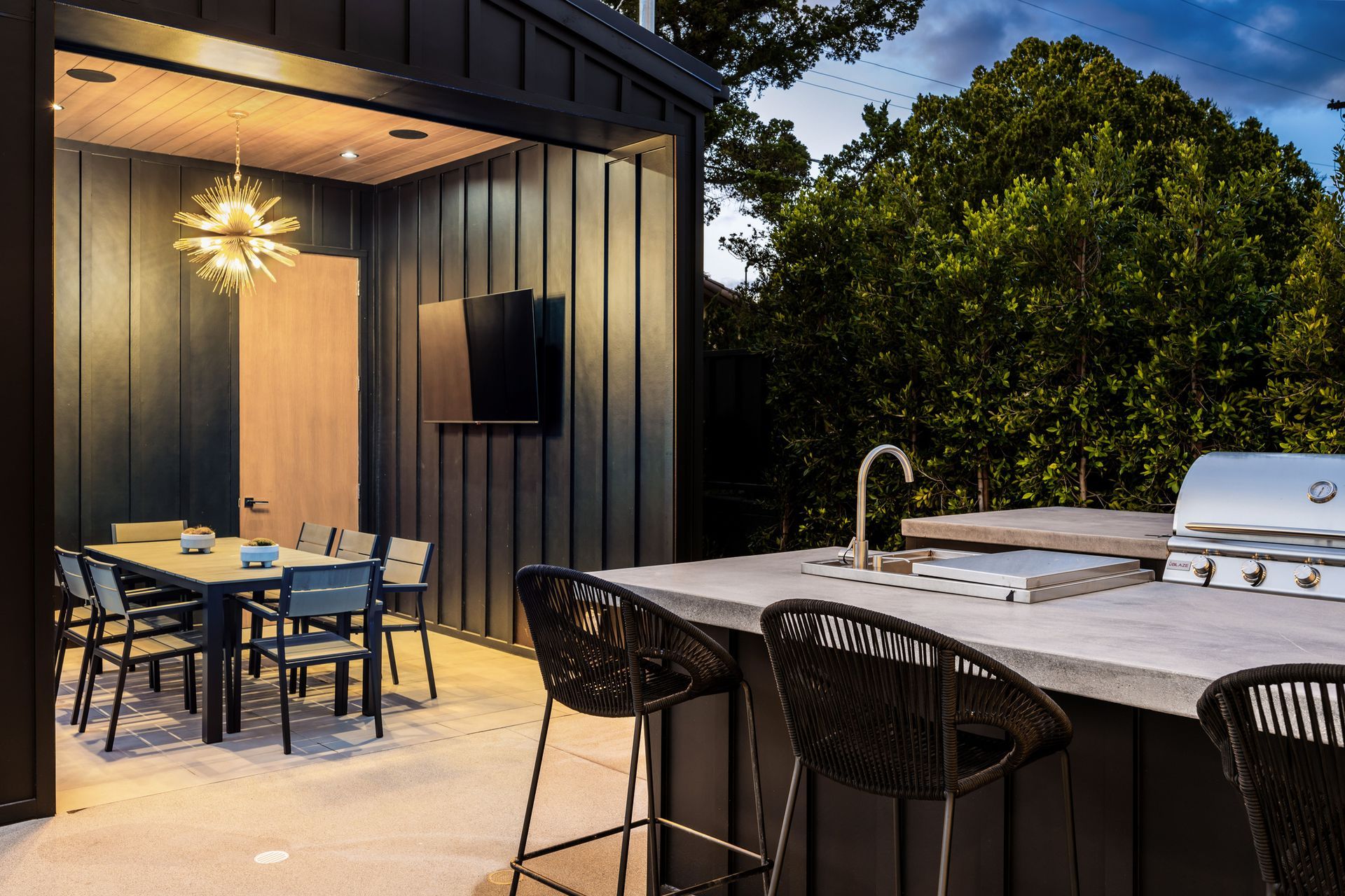 Modern outdoor patio with dining table, bar stools, and a built-in grill beside a lit doorway at dusk