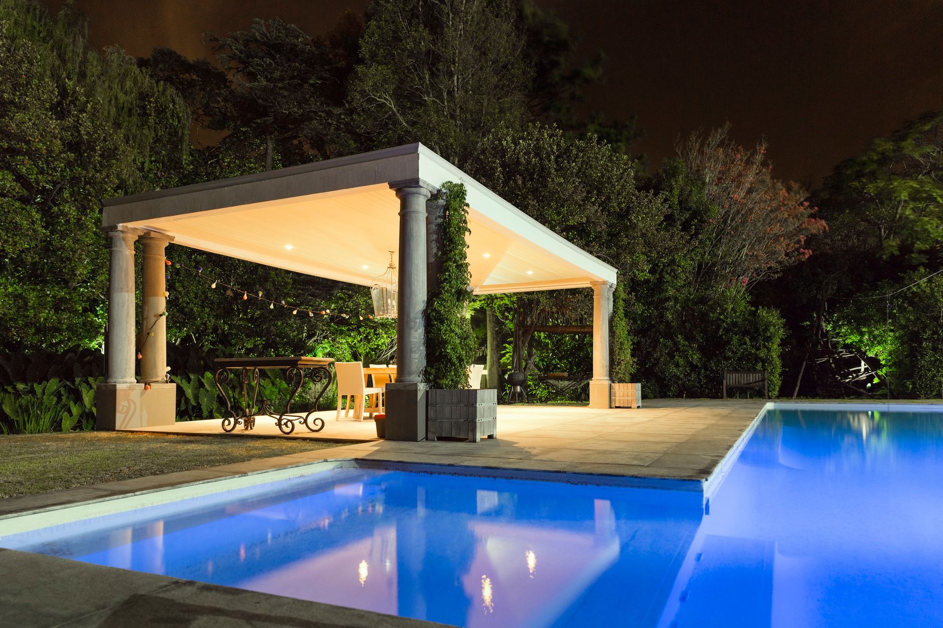 Lit pool beside a white gazebo pavilion at night, surrounded by trees and reflected in the water.