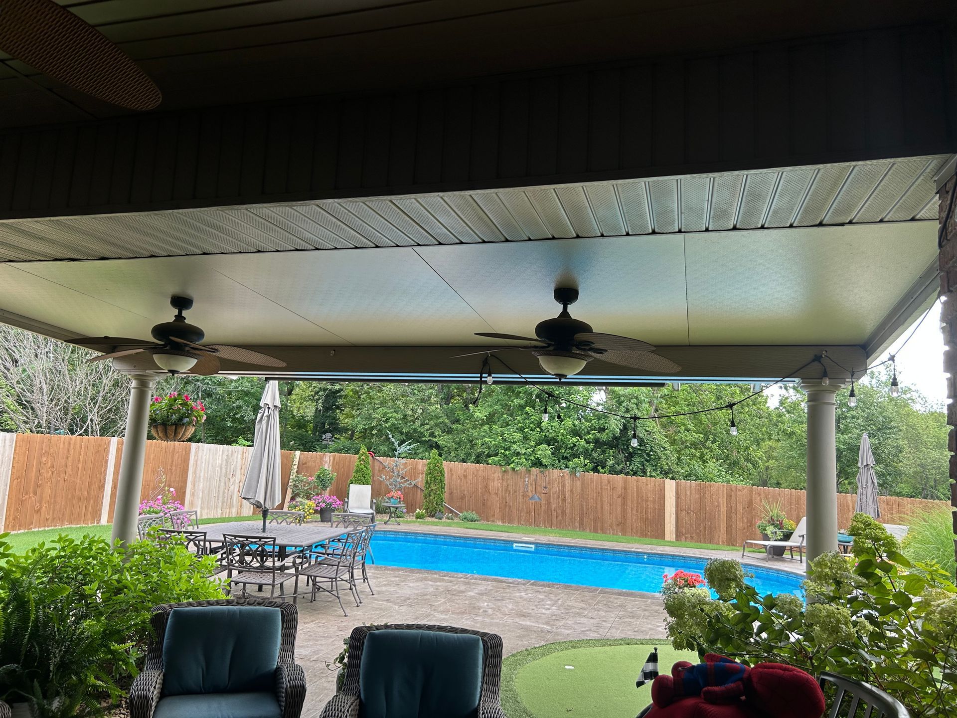 Covered patio overlooking backyard pool and wooden fence, with chairs and ceiling fans.