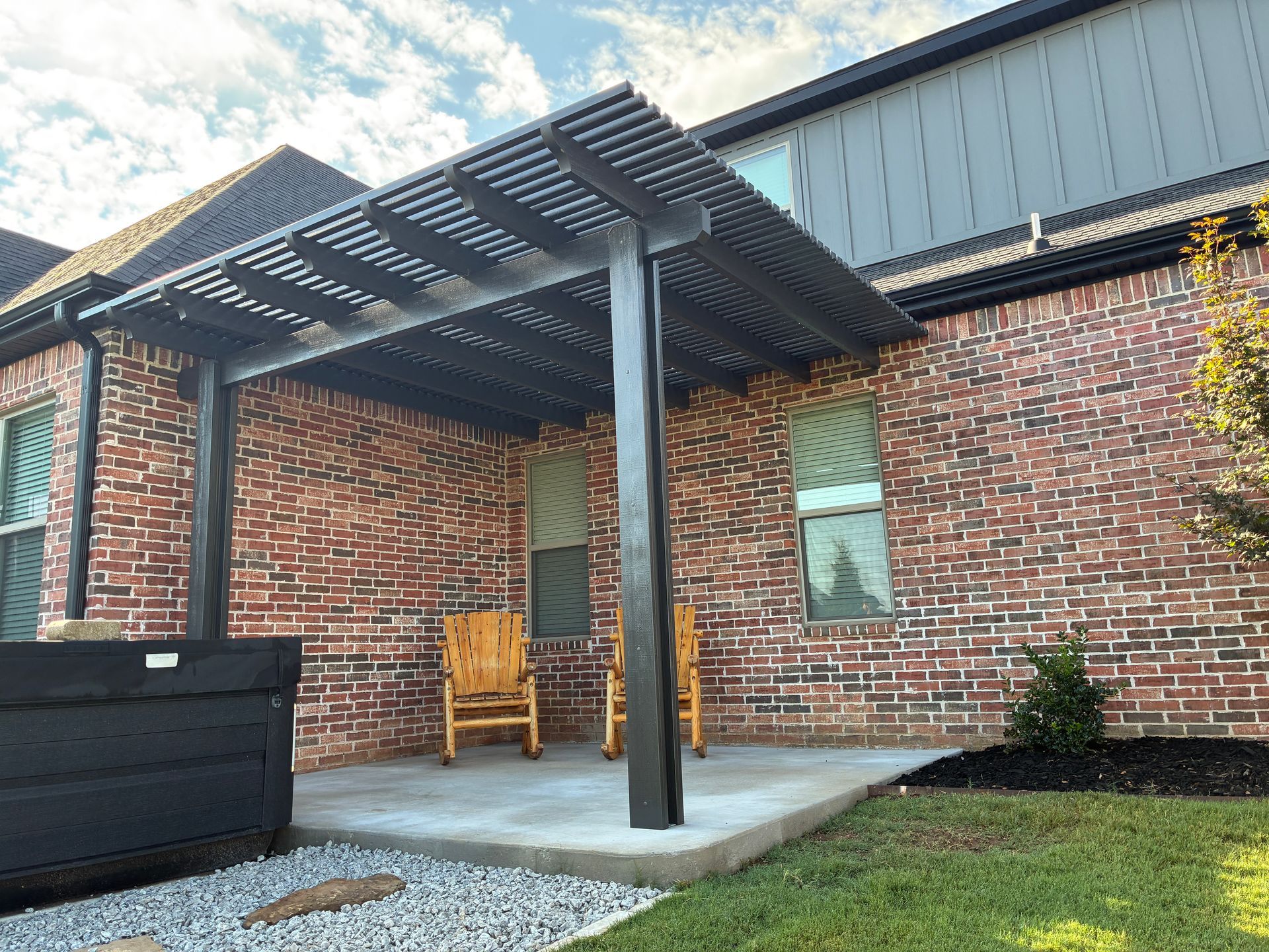 Covered brick patio with metal pergola, concrete slab, and two wooden chairs beside a brick building.