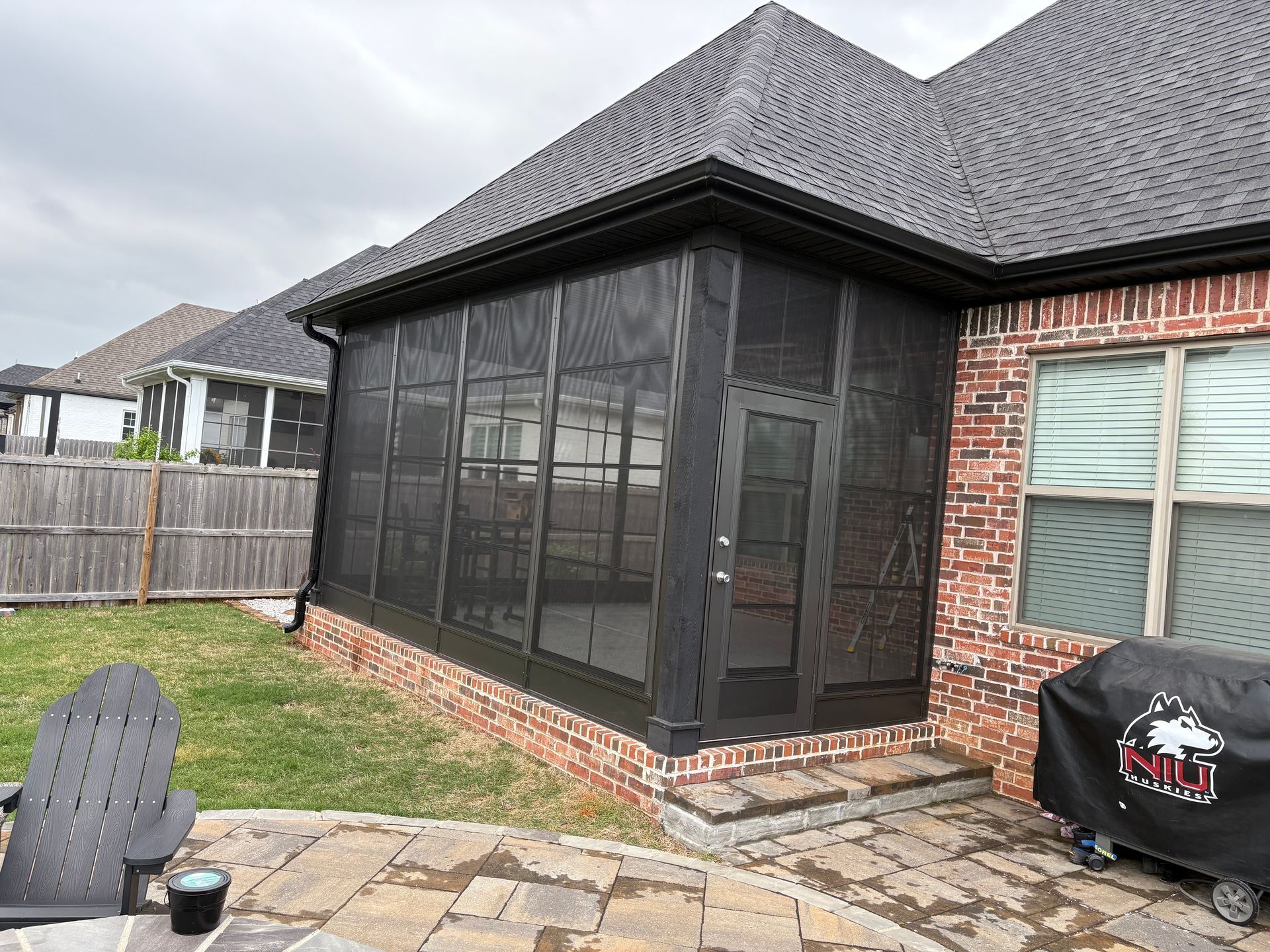 Screened patio attached to a brick house with a black grill and fenced backyard.