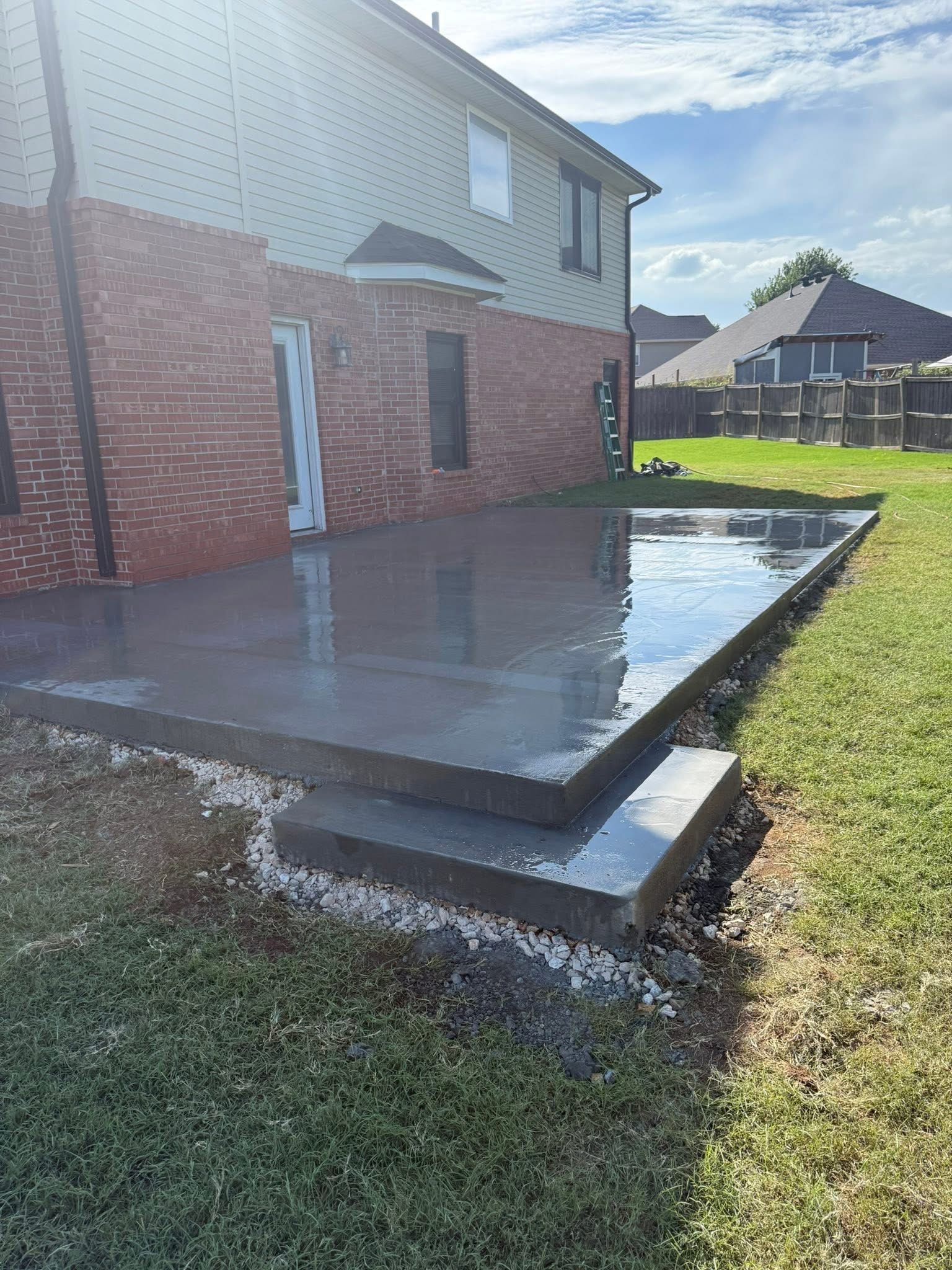 Fresh concrete patio beside a brick house, with a small step and grassy yard under bright sunlight