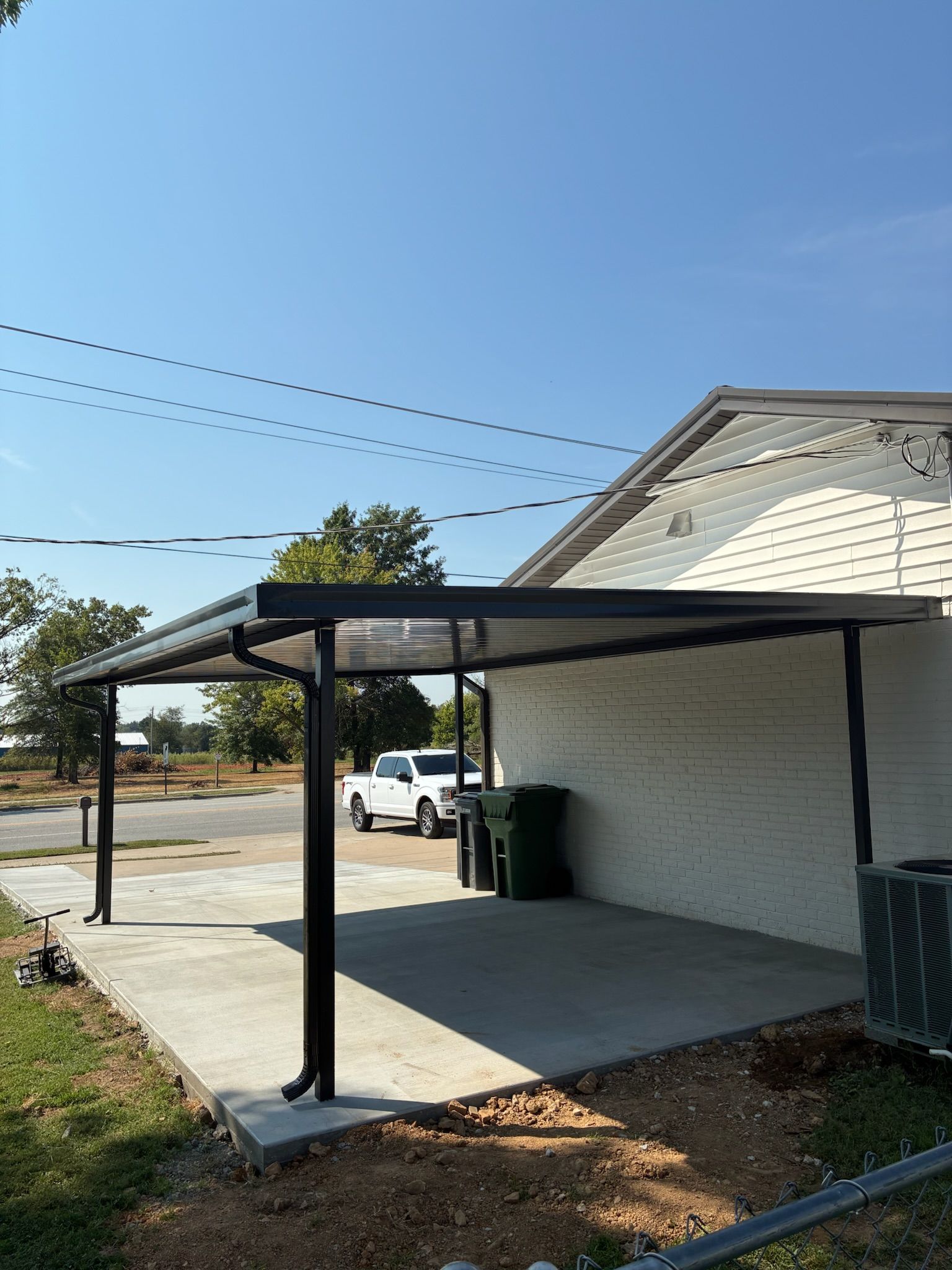 Covered carport attached to a white house with a concrete slab and a trash bin nearby.